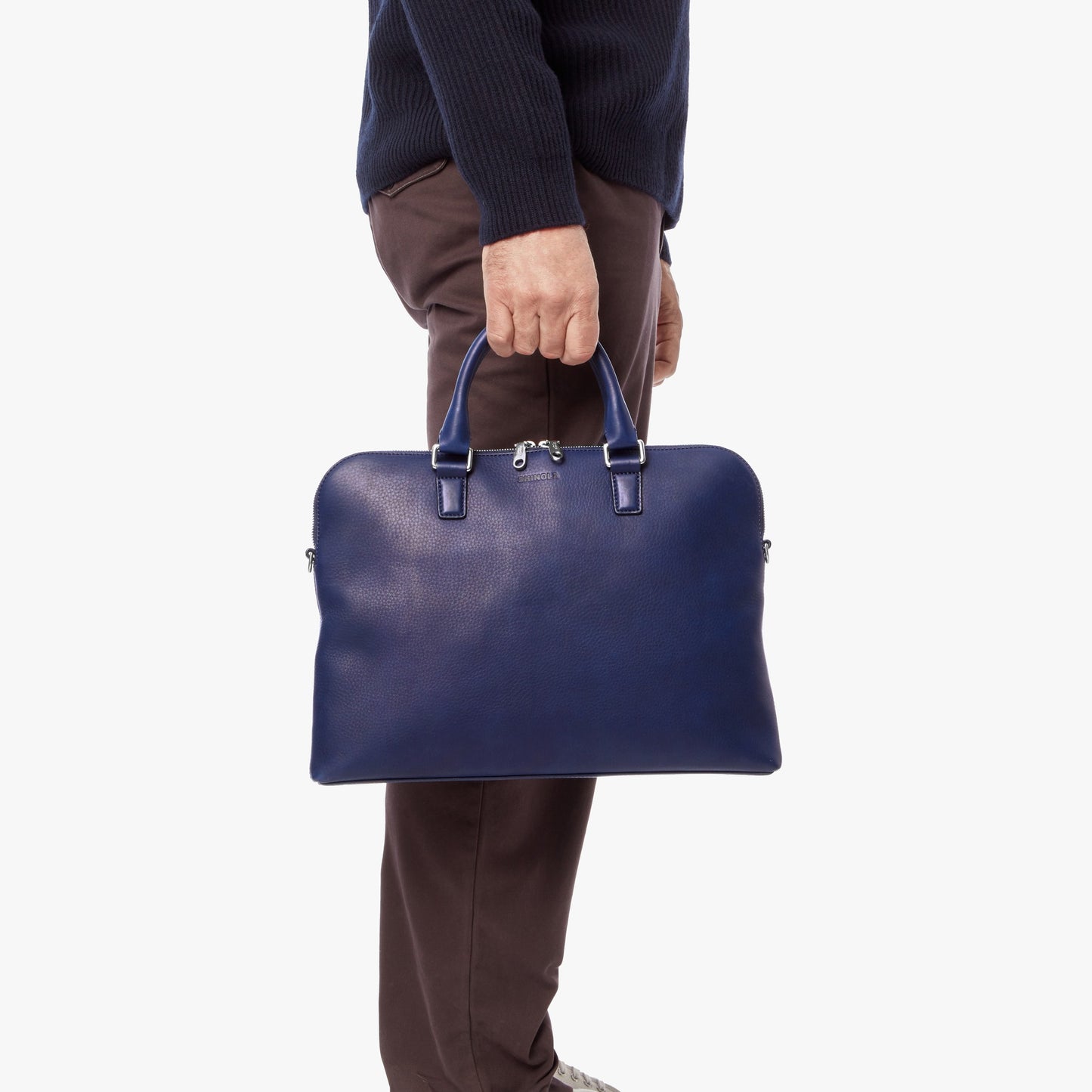 A person in brown pants and a navy sweater holds the Shinola Portfolio Briefcase - Lake, crafted from blue European grain leather, by the handles against a plain white background.