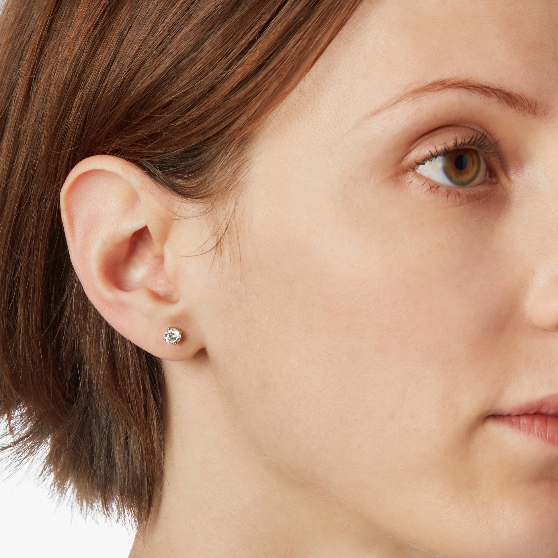 Close-up of a woman with fair skin and short brown hair, wearing Shinola’s 1 Carat Round Diamond Studs in yellow gold. The focus is on her ear and cheek against a plain white background.