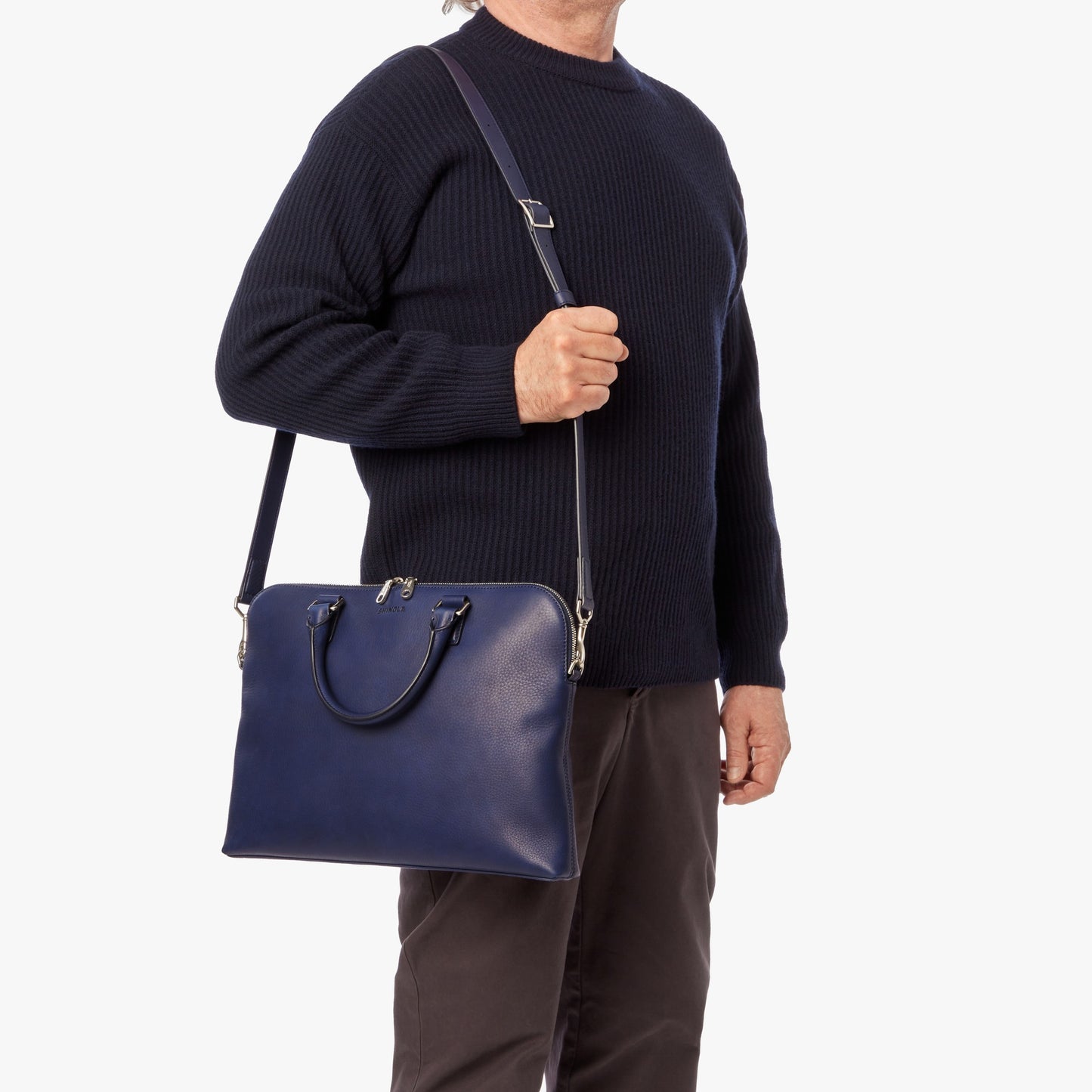 A person dressed in dark attire carries the Shinola Portfolio Briefcase - Lake, a navy blue European grain leather slim laptop briefcase with handles and a shoulder strap, shown against a plain white background.