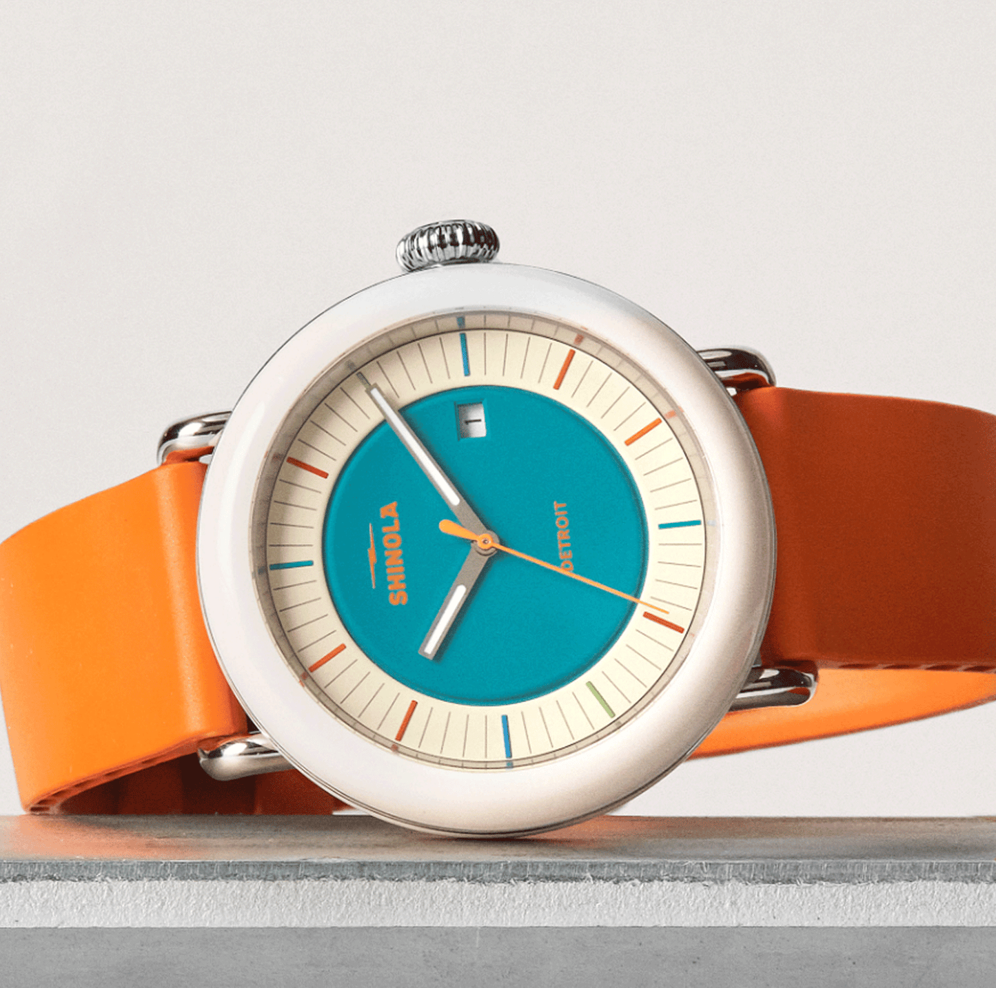 The Shinola Detrola Watch - Aqua Green features a modern art design with an orange band, round silver bezel, blue center dial, cream outer ring with colored markers, and a small date window, shown angled on a gray surface.