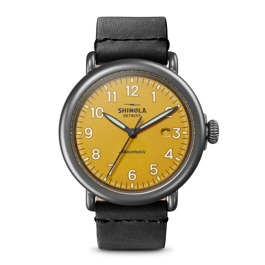 The Shinola Runwell Automatic Watch - Ochre features an ochre vellum-textured dial with black numerals, a date window at 3 o'clock, and a black leather strap. "Shinola" and "Automatic" are printed on the watch face.