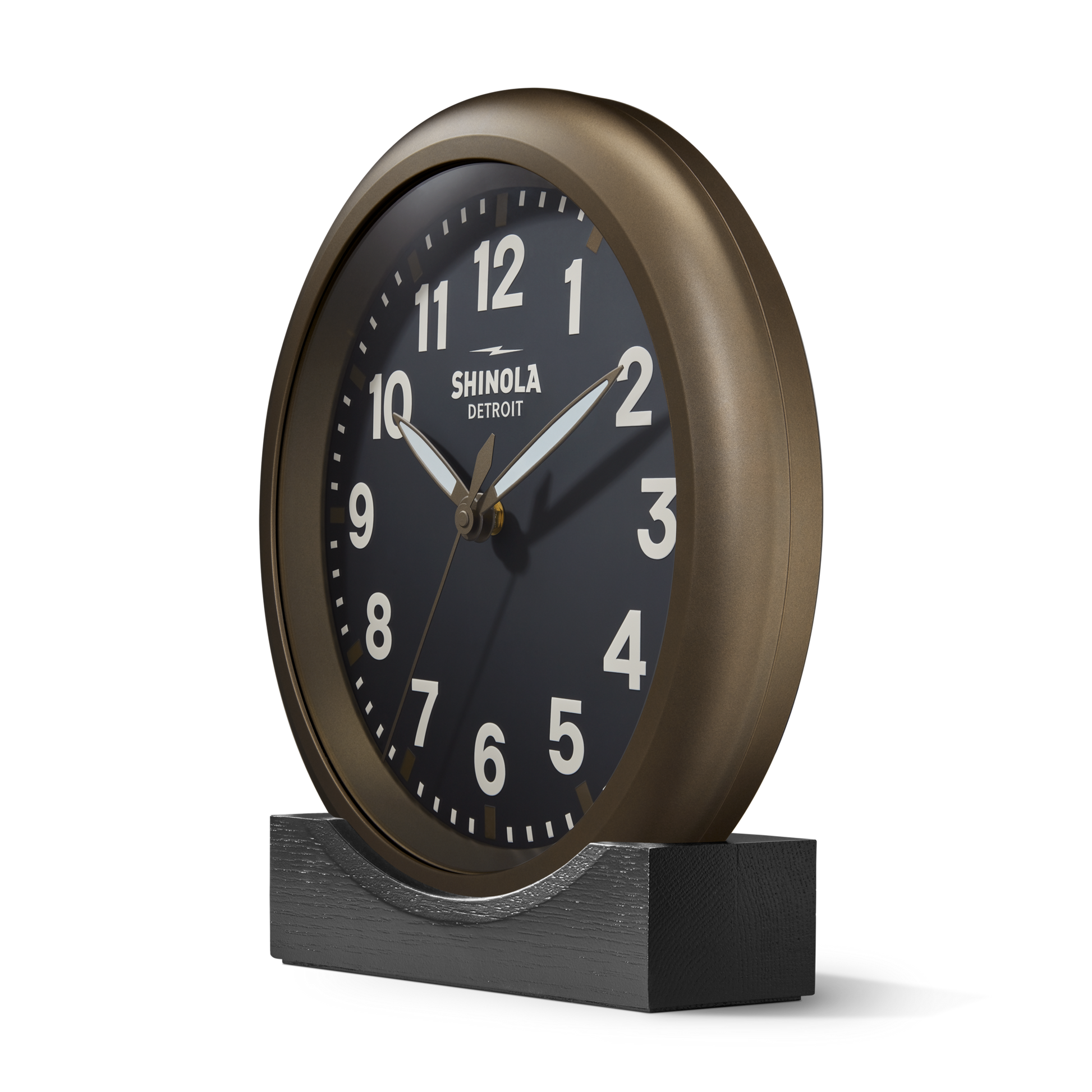 The 14" Runwell Clock - Black by Shinola features a bronze round case, black face with white numerals, and branding. Inspired by the classic Runwell Wall Clock, it sits on a black base and displays the time as 1:10:36.