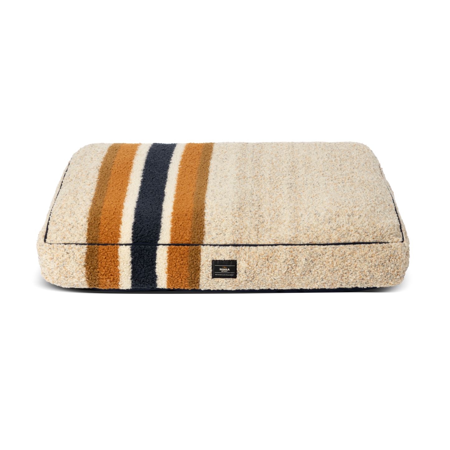The Shinola Napper Pillow Dog Bed - Stripe Brown is a rectangular beige plush bed with memory fiber fill, three horizontal stripes in dark blue, gold, and orange, a small black label, and a machine washable cover. Background: plain white.