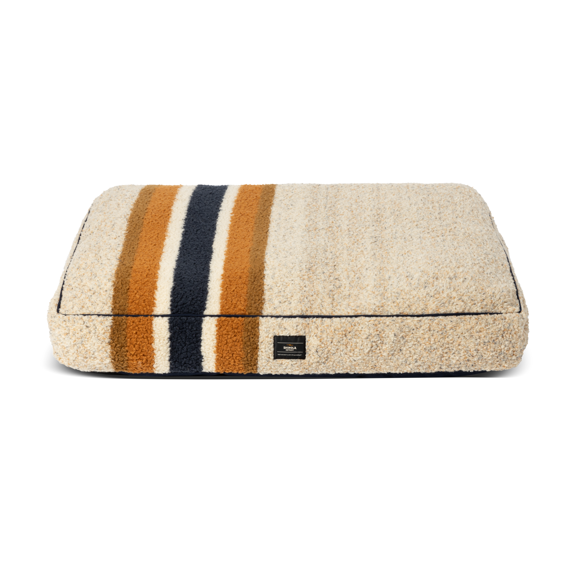 The Shinola Napper Pillow Dog Bed - Stripe Brown is a rectangular beige plush bed with memory fiber fill, three horizontal stripes in dark blue, gold, and orange, a small black label, and a machine washable cover. Background: plain white.