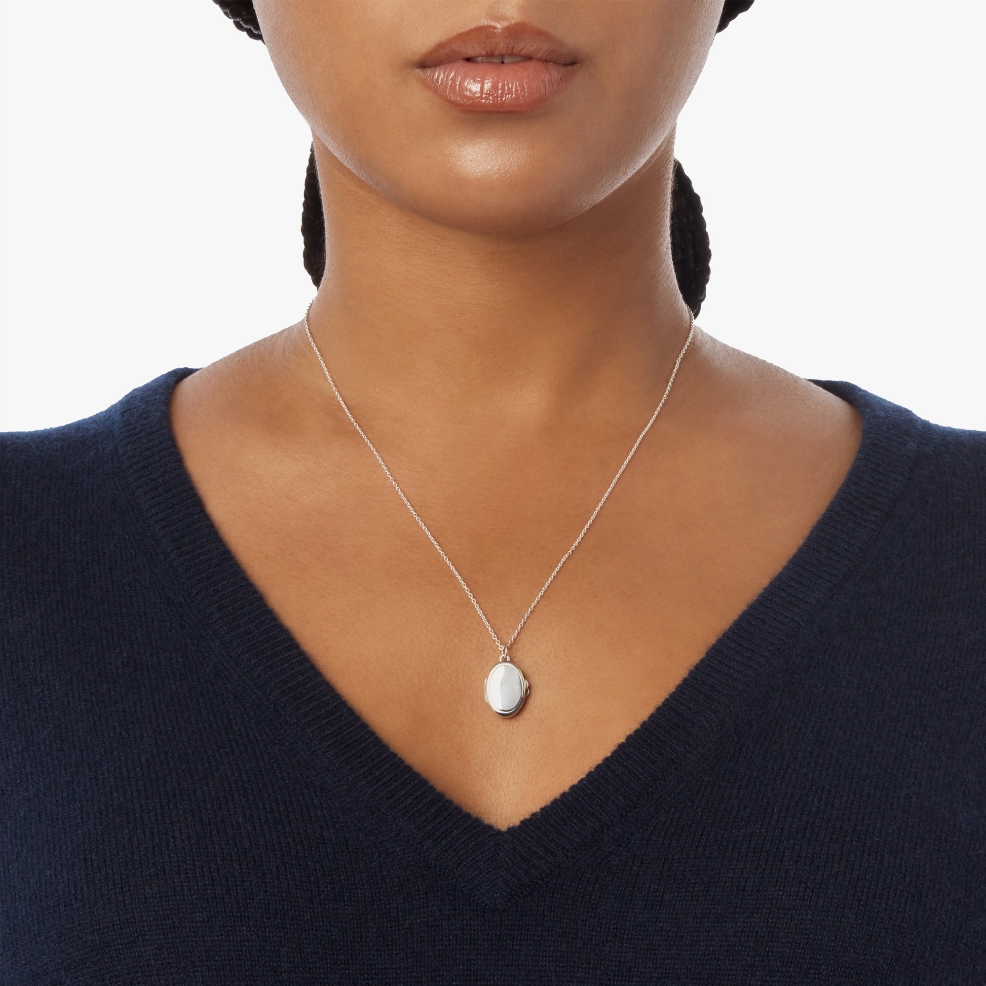 A woman shown from shoulders to just above the lips wears a navy V-neck sweater and the Shinola Heirloom Petite Locket Necklace in sterling silver, featuring a small oval pendant, against a plain white background.