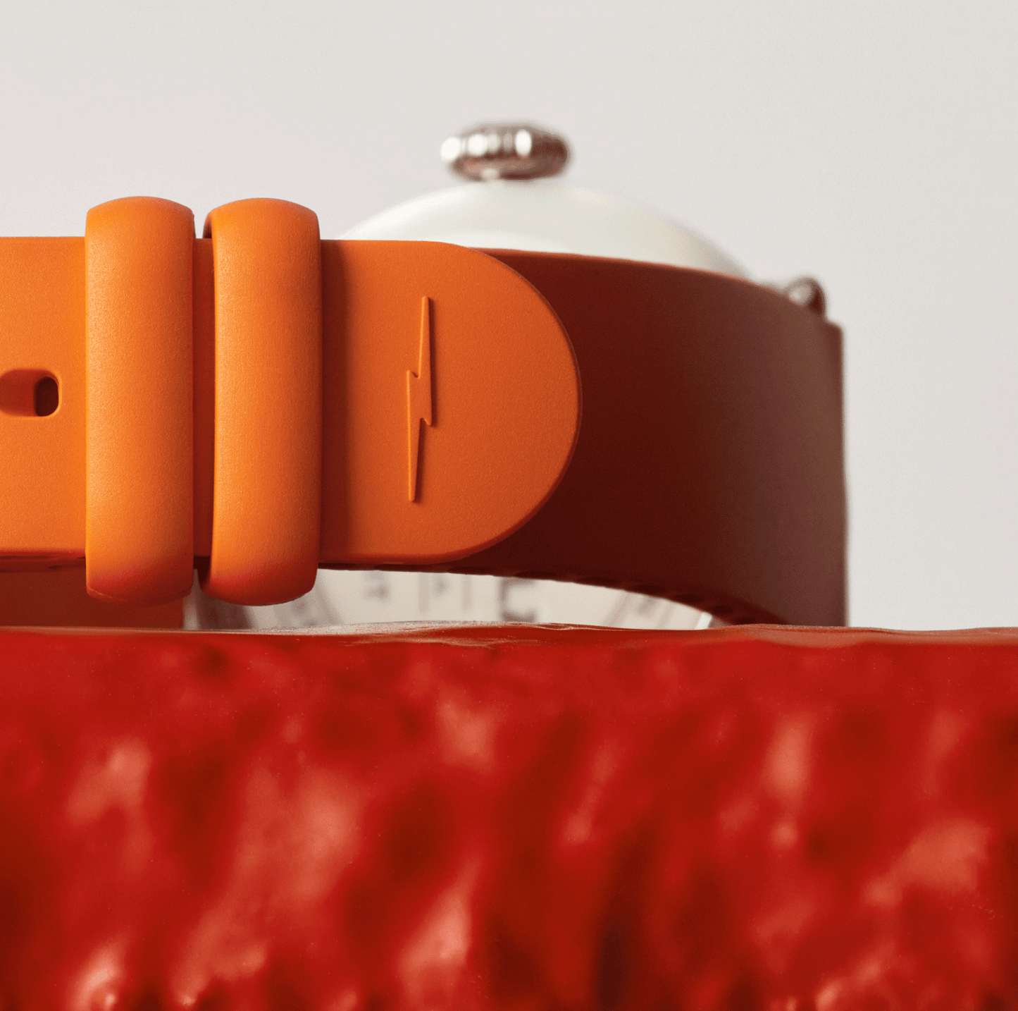 Close-up of an aqua green Shinola Detrola Watch band with a lightning bolt symbol, resting on a textured red surface, with part of the modern art watch face and metallic crown visible in the background.