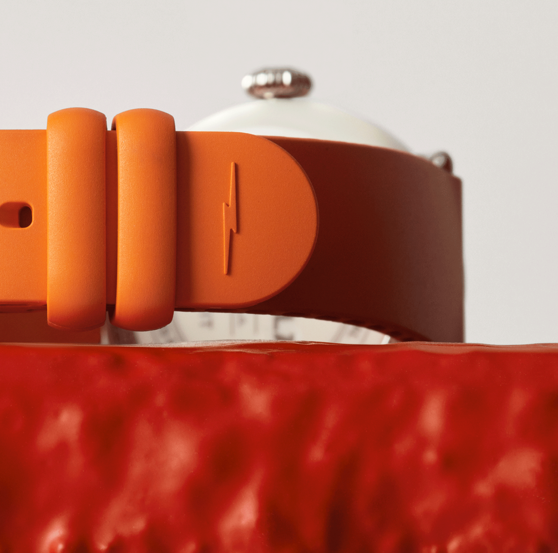 Close-up of an aqua green Shinola Detrola Watch band with a lightning bolt symbol, resting on a textured red surface, with part of the modern art watch face and metallic crown visible in the background.