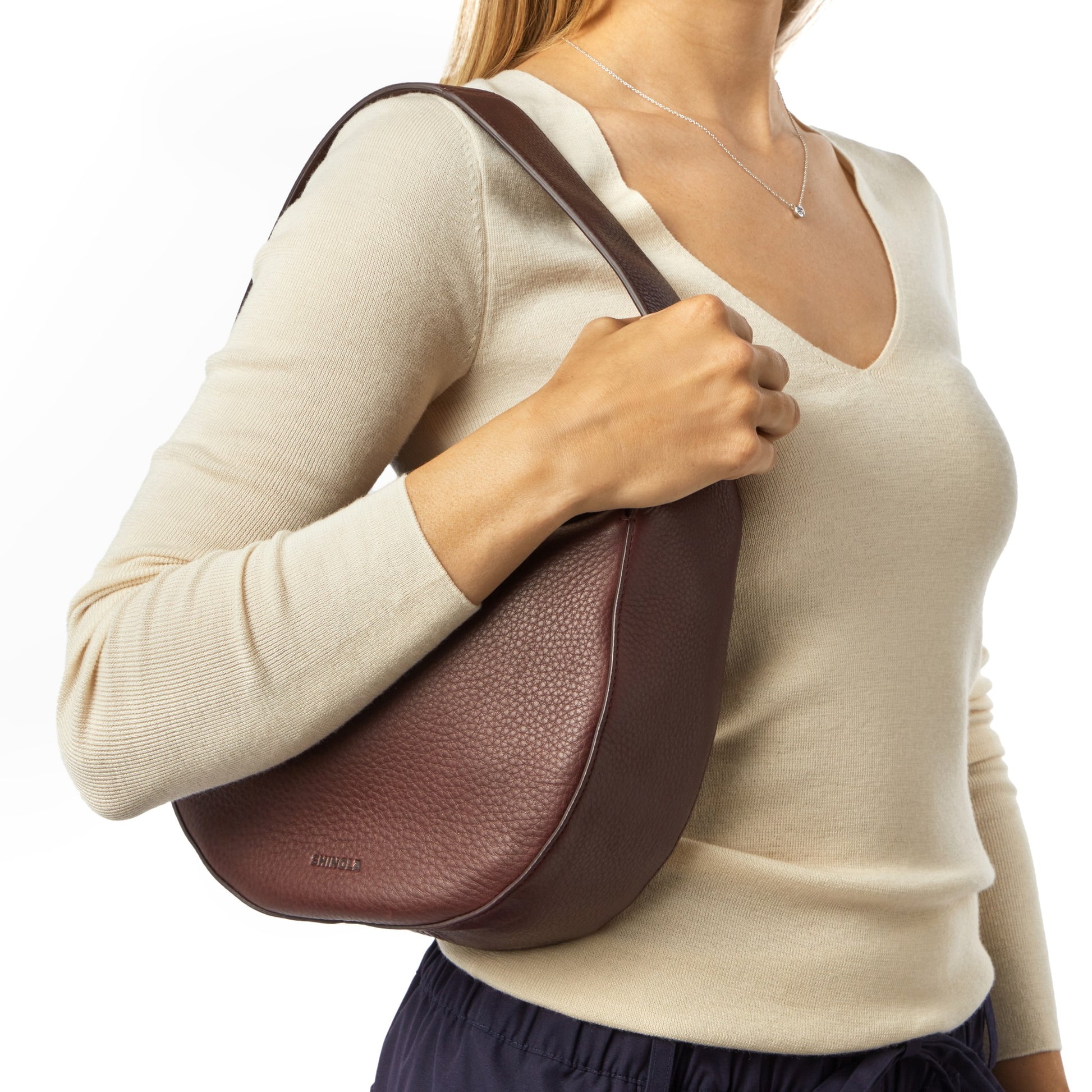 A woman in a cream sweater and navy pants holds the Shinola Small Shoulder Bag - Merlot under her arm. Made from natural grain leather with a textured surface and short strap, only her upper body appears against a white background.