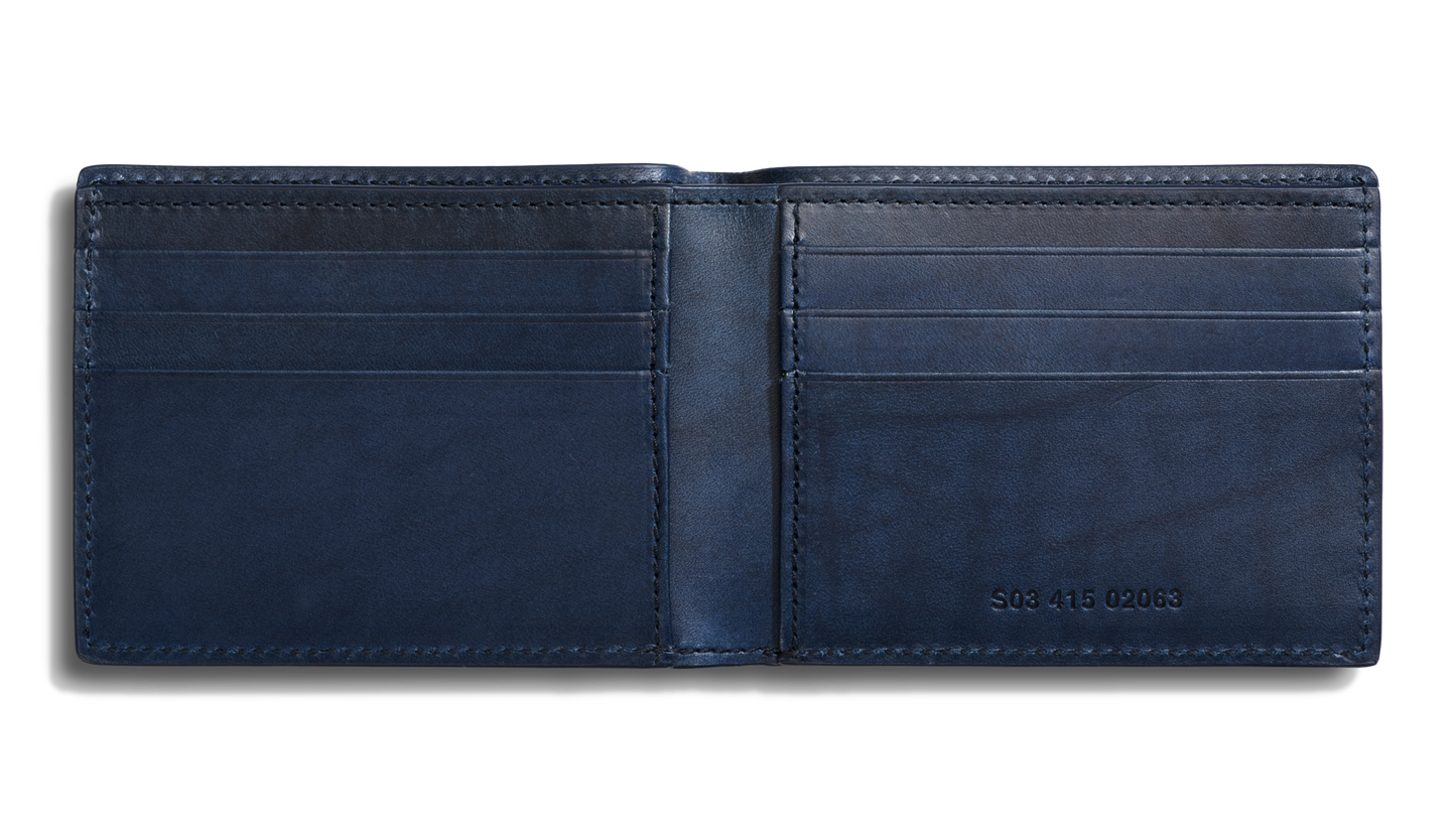 The Shinola Slim Bifold Wallet - Navy is open, revealing card slots on both sides, a bill pocket, and RFID protection. A small set of numbers is embossed on the inside lower right panel.