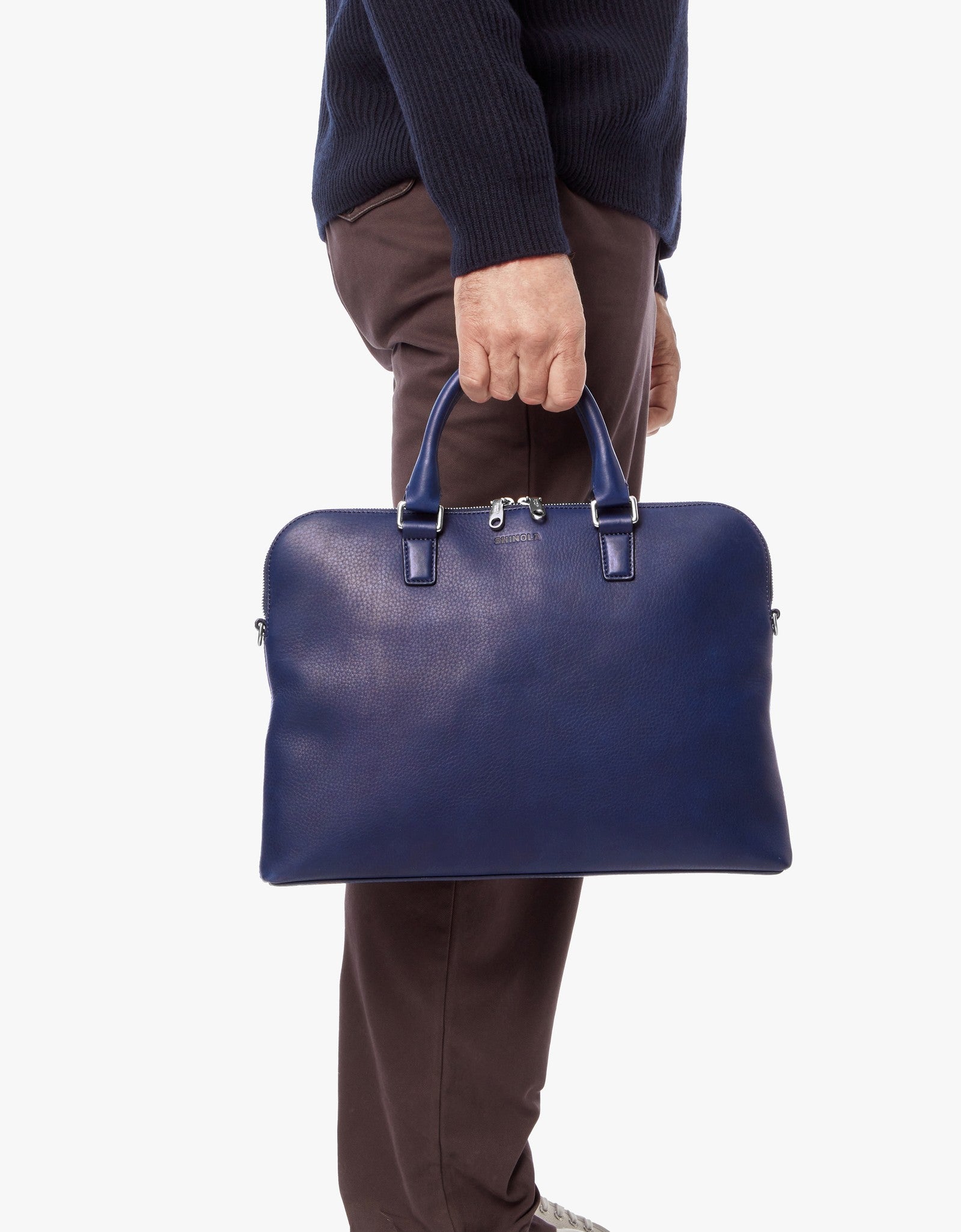 A person in a navy sweater and brown pants holds the Shinola Portfolio Briefcase - Lake, made from European grain leather, by its handles at their side against a plain white background.