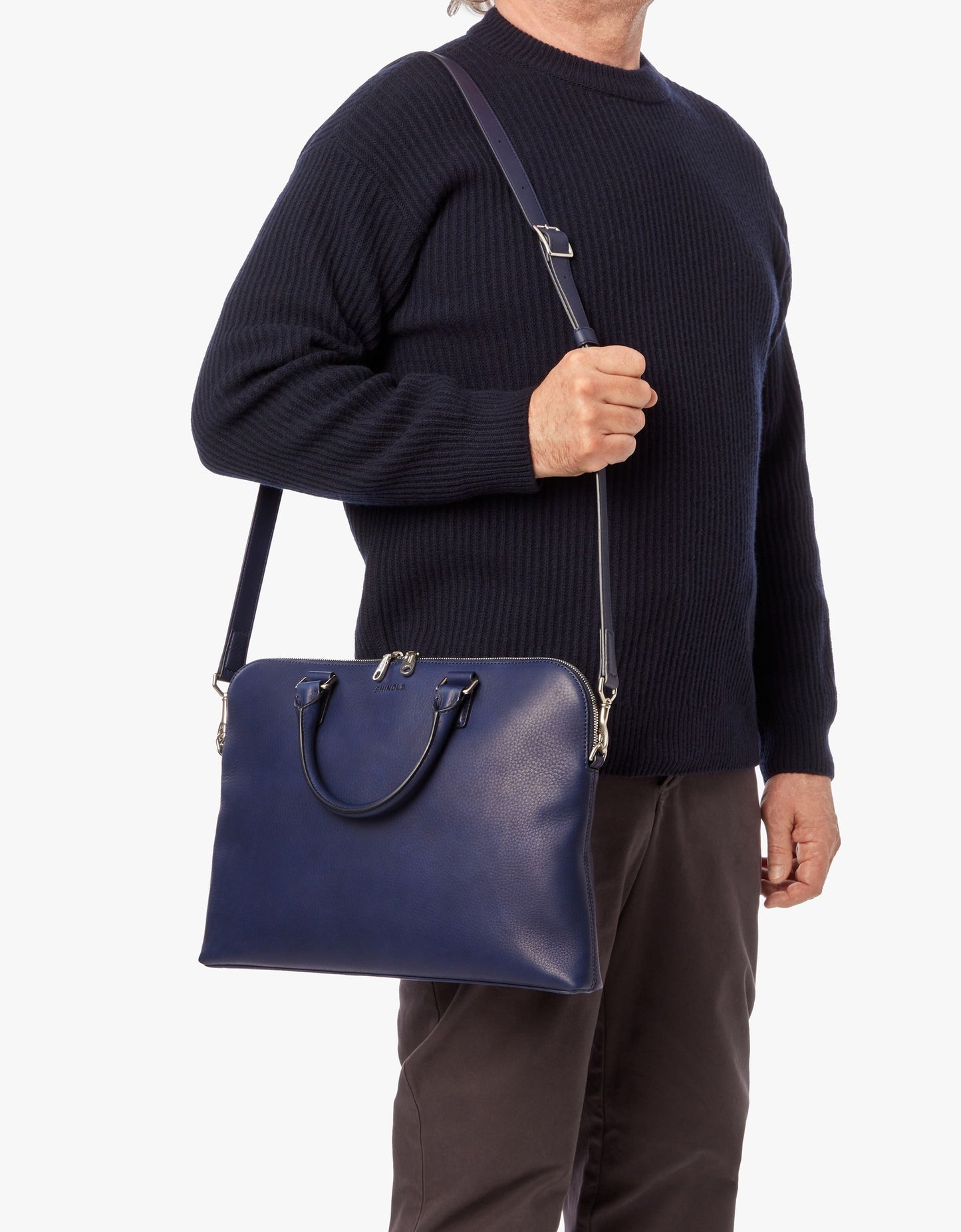 A person in a dark sweater and pants carries the Shinola Portfolio Briefcase - Lake, a navy blue European grain leather laptop bag with handles and an adjustable strap, shown against a plain white background.