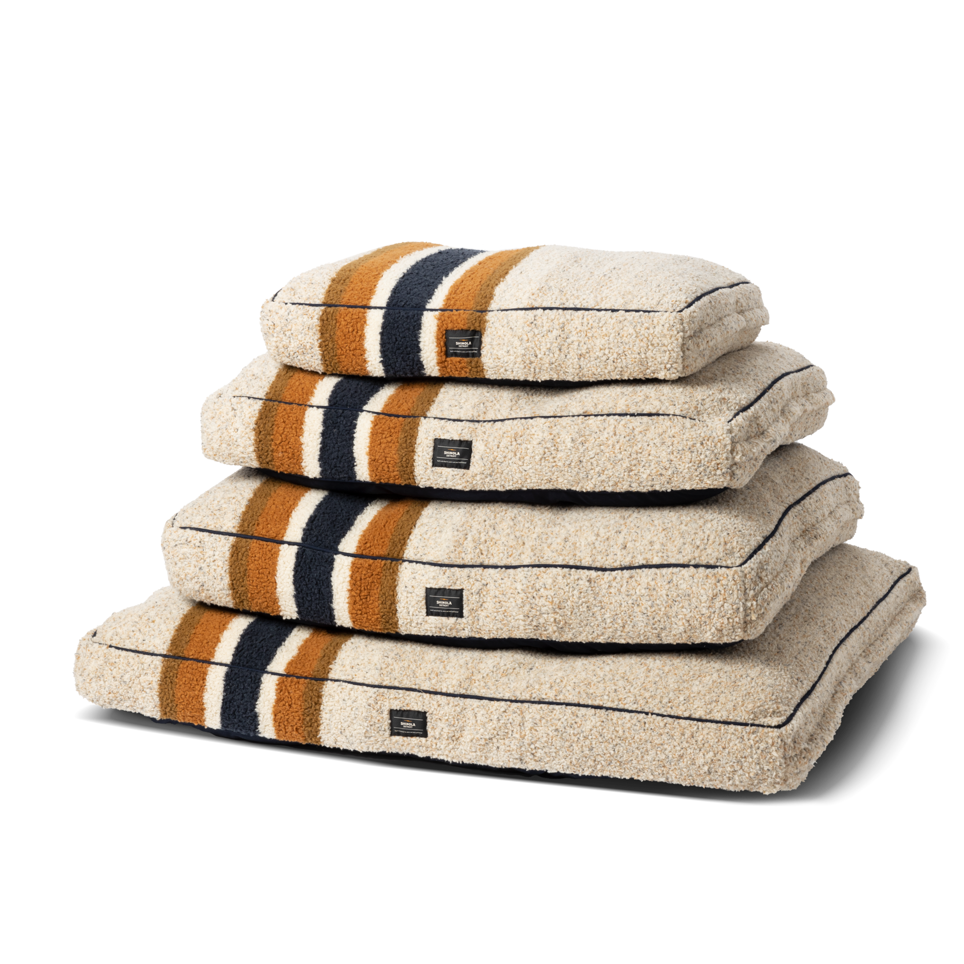 Four Shinola Napper Pillow Dog Beds – Stripe Oat, with a soft shearling Berber cover in brown, orange, and black stripes, are stacked by size against a white background. Each bed is machine washable for easy care.