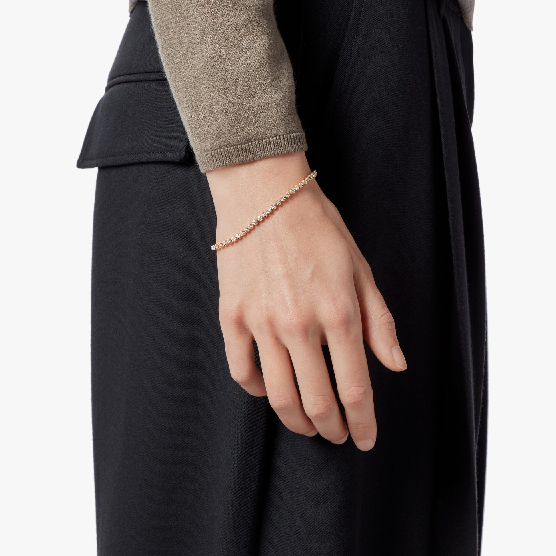A close-up of a hand wearing the Shinola 1.84 Carat 6.5" Diamond Tennis Bracelet in yellow gold, styled with dark high-waisted trousers and a beige long-sleeve top.
