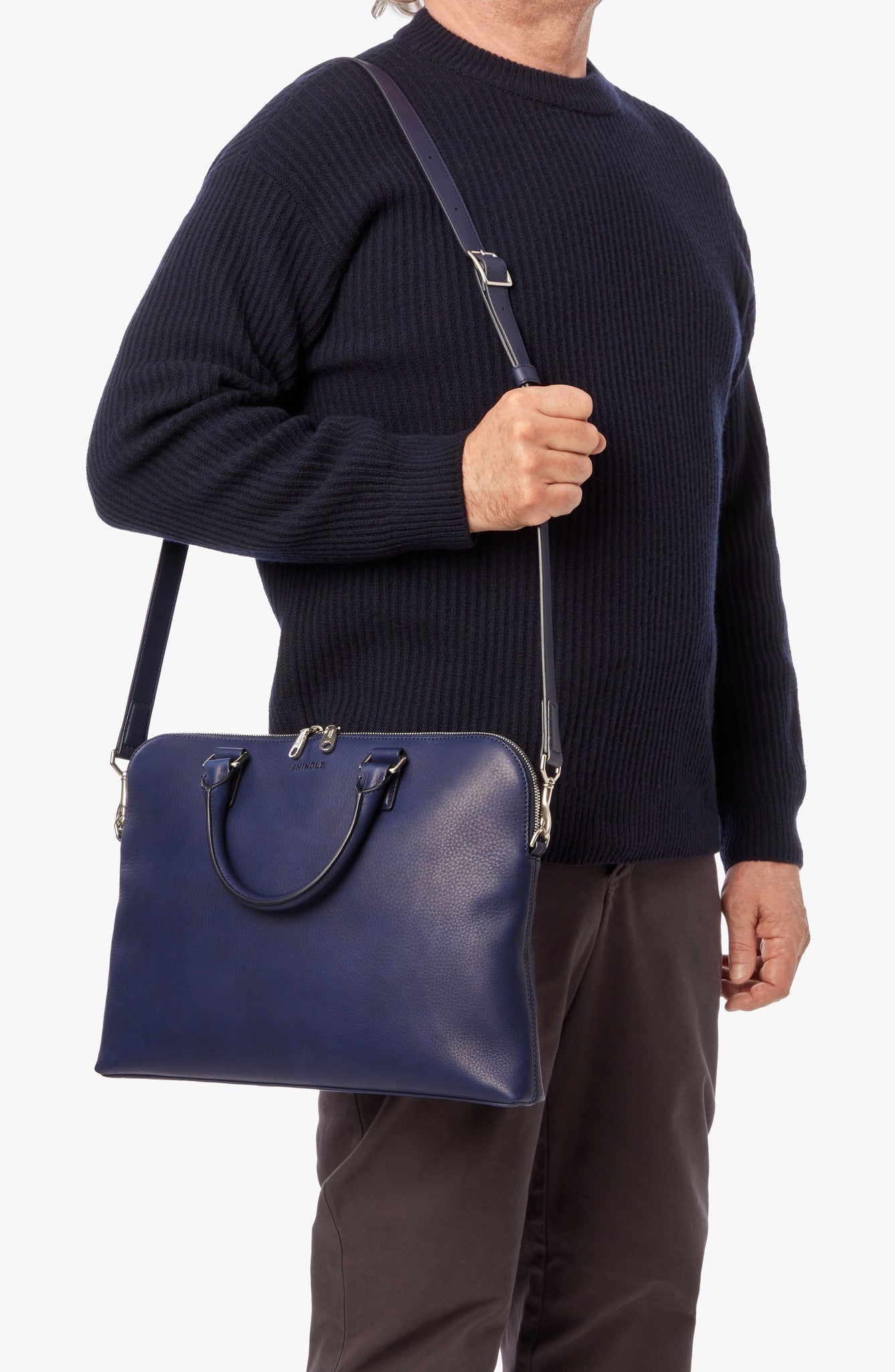 A person in a dark sweater and pants carries the Shinola Portfolio Briefcase - Lake, a navy blue European grain leather slim laptop briefcase with handles, set against a plain white background.