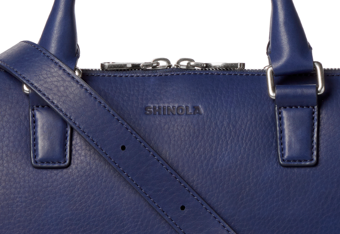 Close-up of a textured blue European grain leather Portfolio Briefcase - Lake by Shinola, featuring silver hardware, an embossed logo on the front, and a shoulder strap.