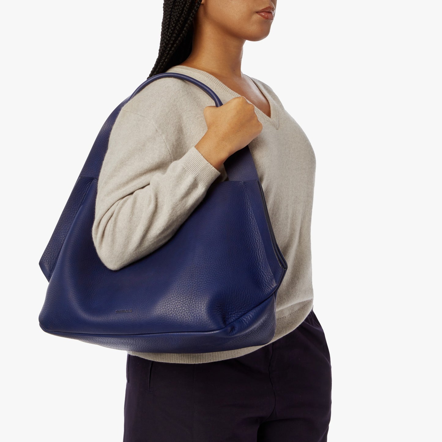 A woman in a light beige sweater and dark pants carries the Shinola Runwell Shoulder Bag - Lake with a leather card pocket on her right shoulder against a plain white background.