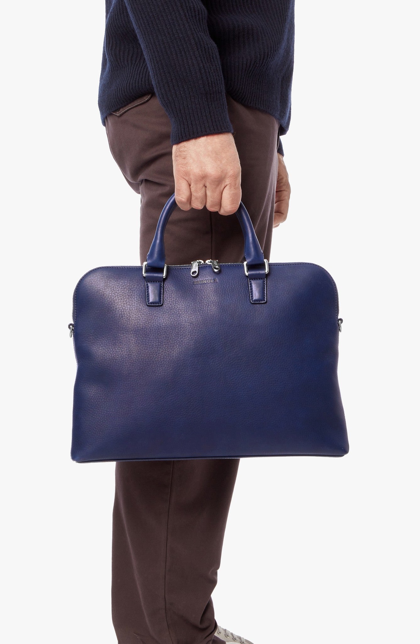 A person wearing a dark sweater and brown pants holds the Shinola Portfolio Briefcase - Lake, crafted from European grain leather, by its handles against a white background.