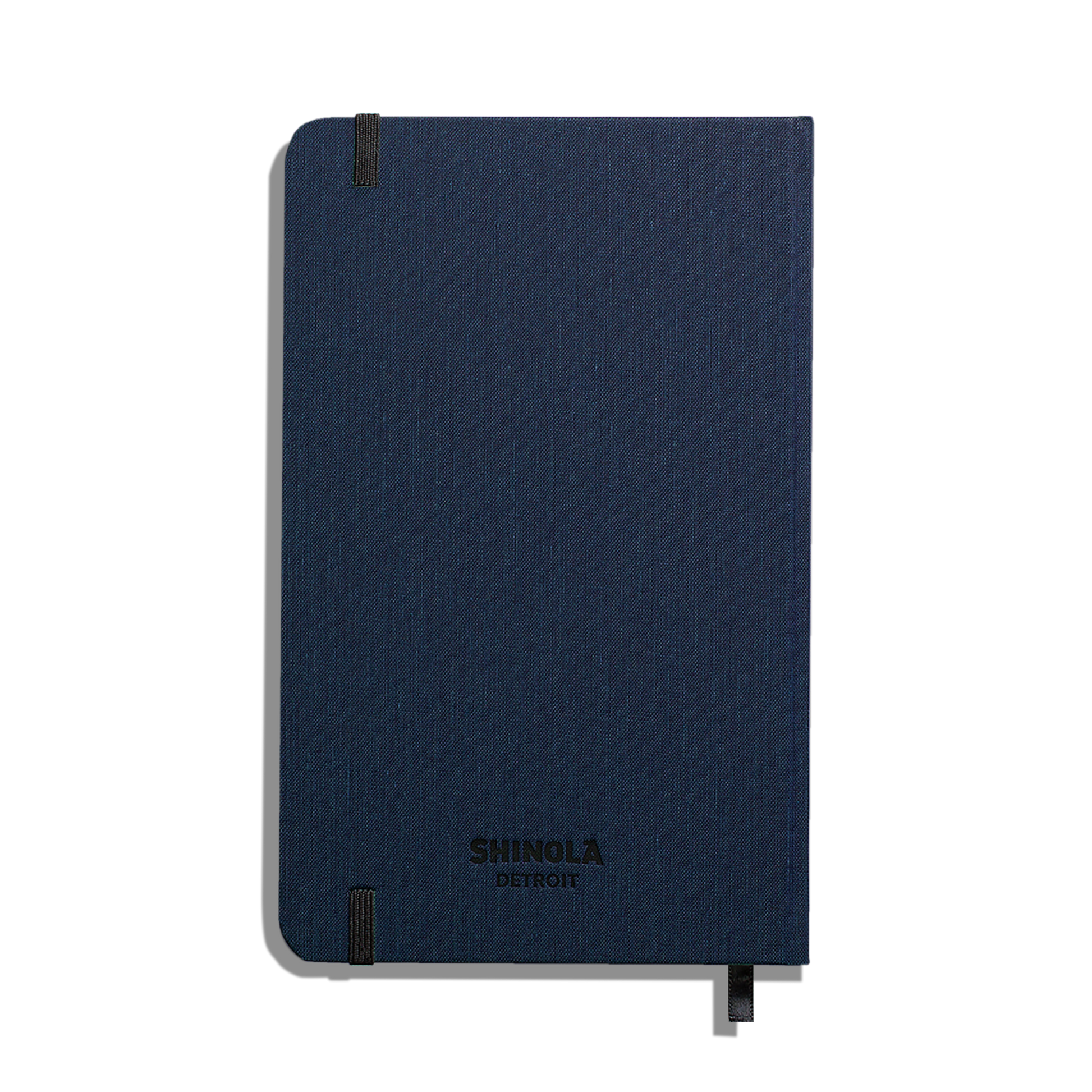 A closed Shinola Medium Hard Ruled Journal in navy with black elastic bands at two corners, smyth-sewn binding, and a matching ribbon bookmark is shown against a plain white background.