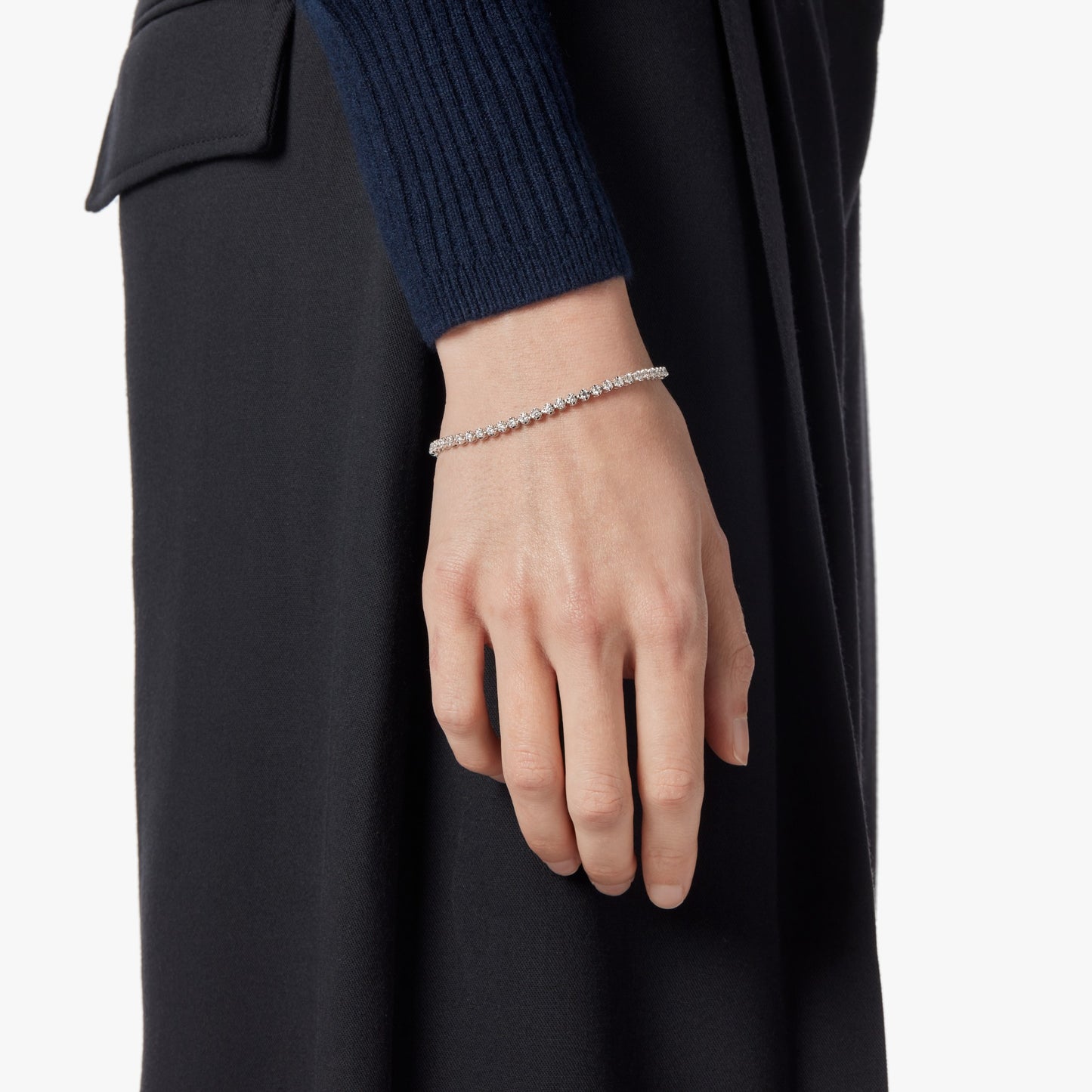 A person in a dark long-sleeve top and black pants is shown from the wrist down, showcasing the Shinola 1.84 Carat 6.5" Diamond Tennis Bracelet in white gold on their left wrist against a plain background.