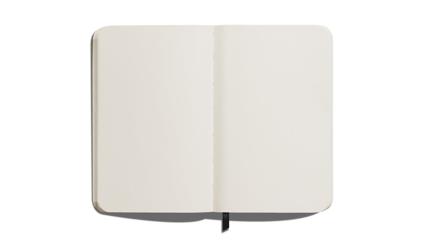 The Shinola Medium Hard Plain Journal in Navy features cream-colored pages, rounded corners, and lies flat on a white surface. A black ribbon bookmark is visible at the bottom center.