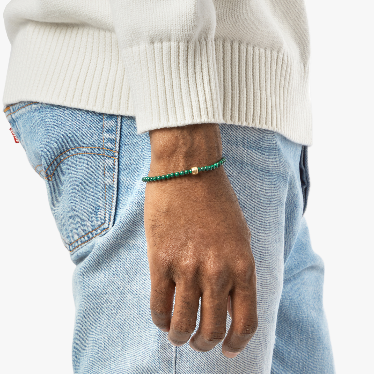 A person in a white sweater and light blue jeans displays their left arm, wearing the Shinola 4mm Beaded Bracelet Malachite. The bracelet features malachite beads and a brass accent bead. The background is plain white.