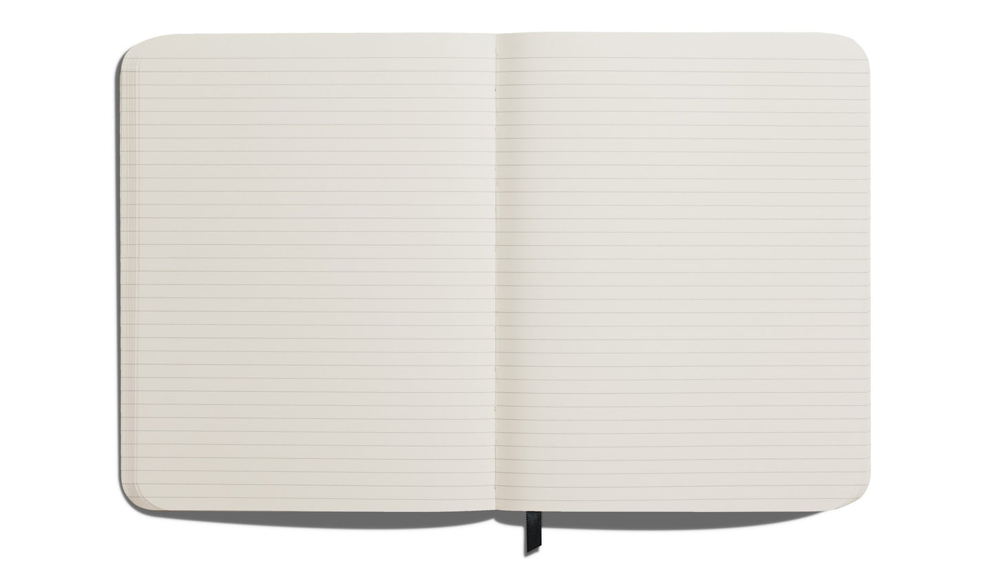 The Shinola Large Hard Ruled Journal in black lies open on a white background, showing its acid-free lined pages and a black ribbon bookmark at the center.
