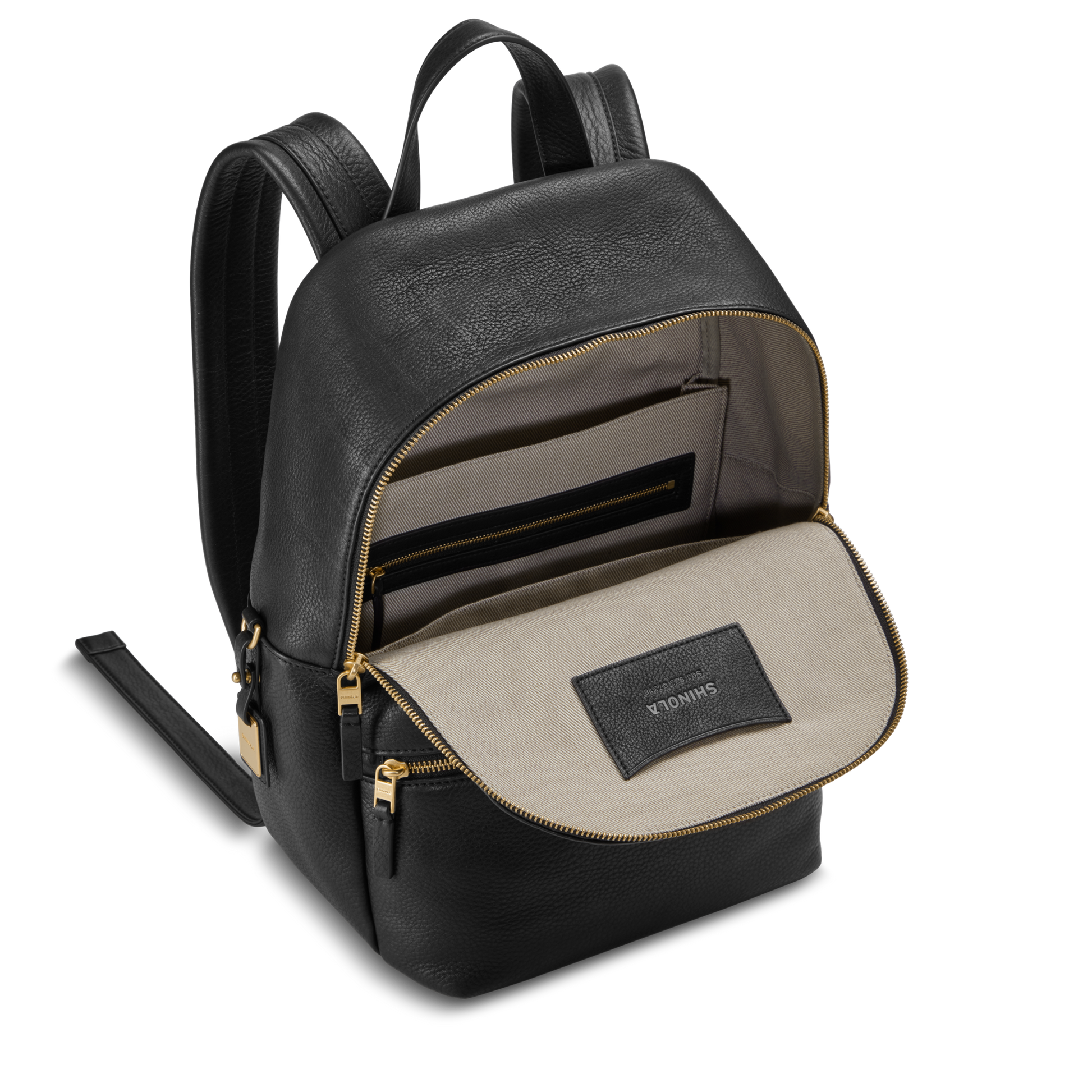 The Small Runwell Backpack by Shinola is shown open in black natural grain leather with gold zippers, revealing a beige fabric interior with a zip pocket and a stitched SHINOLA label.