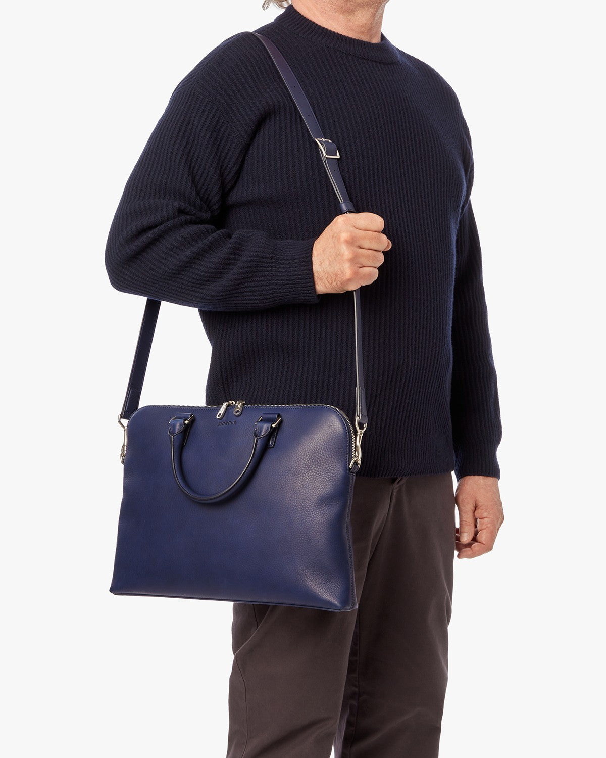 A person in a navy sweater and dark pants holds the Shinola Portfolio Briefcase - Lake, crafted from European grain leather with both handles and a shoulder strap, shown against a plain white background.