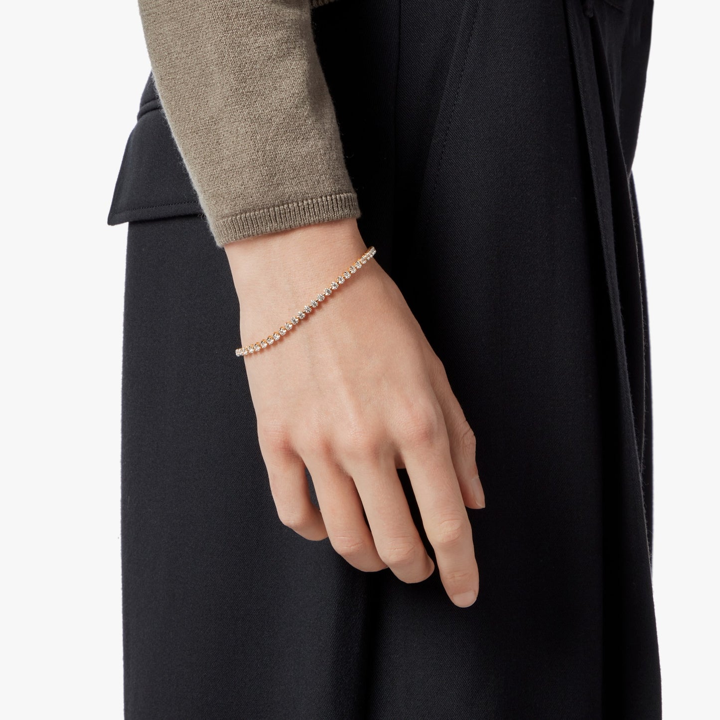 A person in a tan long-sleeve top and black pants wears the Shinola 2 Carat 7" Diamond Tennis Bracelet in yellow gold. Their relaxed hand rests at their side, set against a crisp white background.