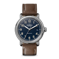 The Shinola Runwell Automatic Watch features a blue dial with silver numerals and hands, a brown leather strap, and a date window at 3 o’clock. The watch face displays "Automatic," showcasing this refined timepiece.